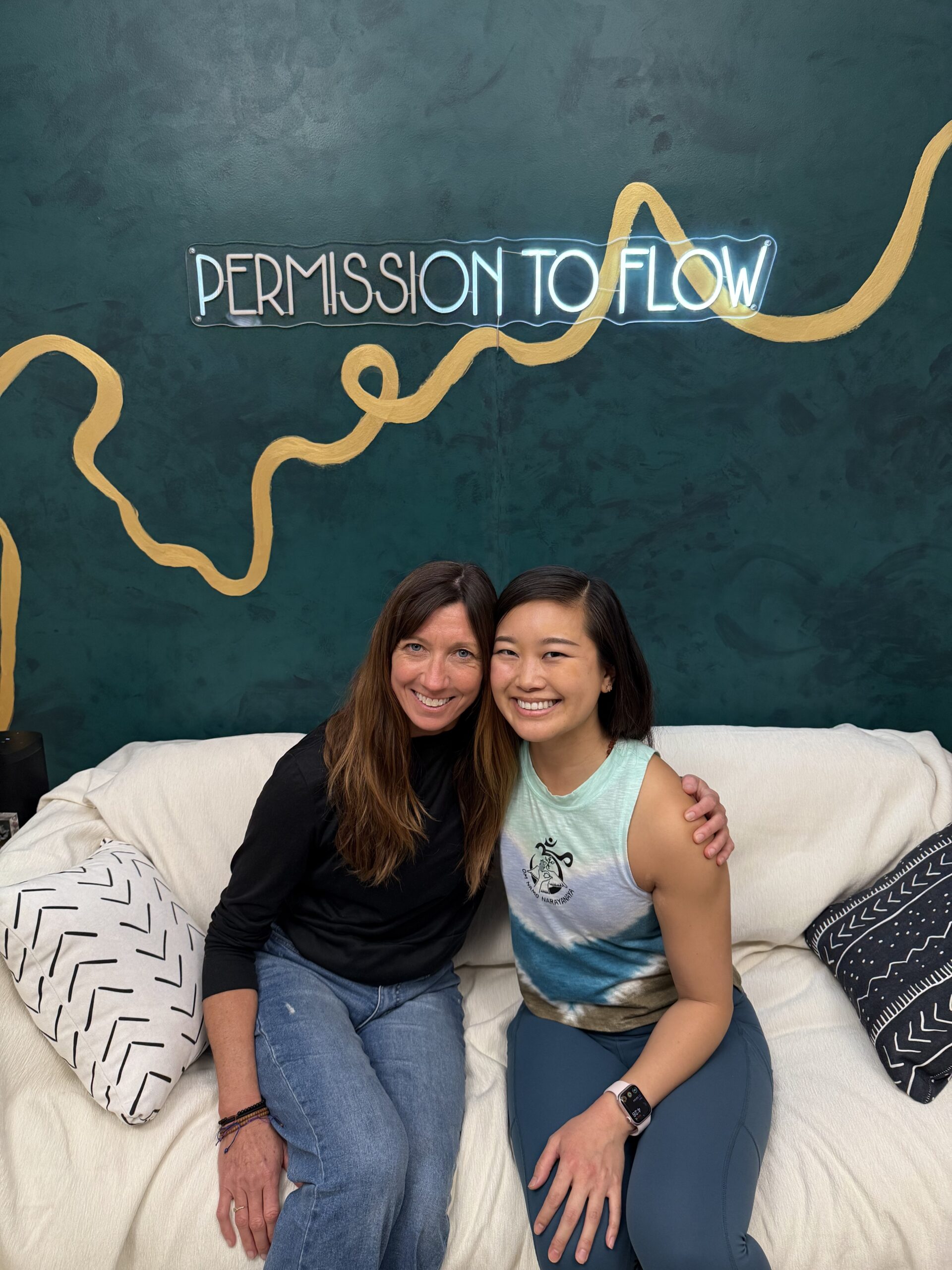 Gabrielle Kali Om and Jessica Hwang sitting on a white couch in front of the permission to flow podcast sign